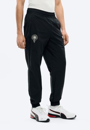 Homme portant un pantalon de jogging noir avec un logo sur la cuisse gauche, des chaussettes blanches, des baskets blanches et noires avec des détails rouges, et un haut sans manches blanc.