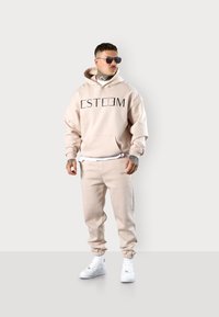 Beige Kapuzenpullover mit "ESTEEM" Druck, passende Jogginghosen und weißen Sneakern. Lockere Passform, weicher Stoff, gerippte Bündchen und Kordelzug an der Kapuze.
