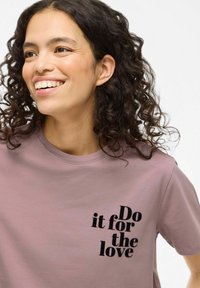 Camiseta de algodón rosa claro con un eslogan en texto negro "Hazlo por amor" en diferentes tipografías en la parte frontal. Diseño de manga corta y cuello redondo.