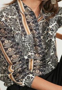 Langärmliges Hemd mit Knopfleiste, das ein schwarz-weißes Blumenmuster zeigt, mit braunen und beigen gestreiften Akzenten sowie einer Brusttasche.