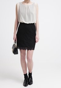 Femme portant un top sans manches blanc, une jupe en dentelle noire, des chaussettes noires et des chaussures noires brillantes, tenant un sac à main noir avec une chaîne dorée.