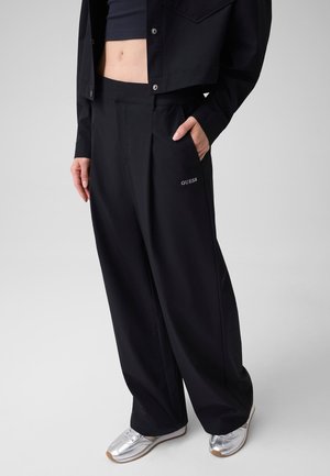 Femme portant un pantalon noir à jambes larges avec le logo "GUESS" sur la poche, une veste courte et des baskets argentées, debout avec une main dans la poche.