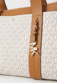 Sac fourre-tout avec un tissu à motifs beige portant les initiales "MK," complété par des anses texturées marron et un pendentif en métal doré.
