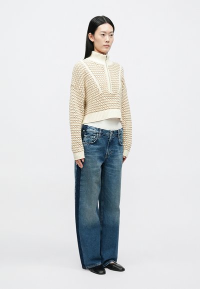 Pulover bej tricotat crop, cu guler înalt și fermoar, asortat cu blugi evazați de culoare albastru deschis. Pantofi slip-on negri completează ținuta.