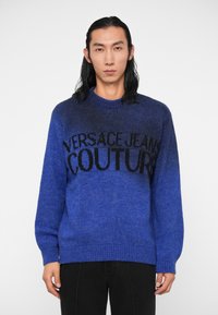 Kék kötött pulóver, amely folyamatos színátmenetet mutat, kerek nyakkivágással és fekete "VERSACE JEANS COUTURE" logóval a mellrészen. Puha tapintású.