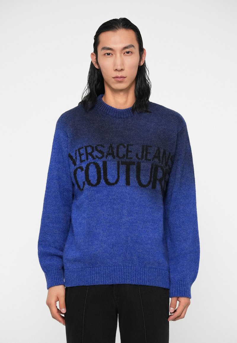 Modra pletena jopica z gradientnim dizajnom, okroglo izrezano ovratnikom in črnim logotipom "VERSACE JEANS COUTURE" čez prsi. Mehka tekstura.