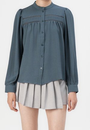 Kvinde iført en langærmet teal skjorte med knapper og blonde detaljer samt en lysegrå plisseret mininederdel, stående foran en hvid baggrund.