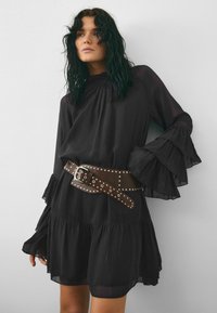 Rochie neagră, cu mâneci lungi, având volane stratificate și guler înalt, asortată cu un cureaua lată din piele, cu ținte maronii. Material neted, ușor.