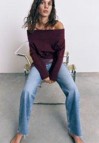 Ung kvinde med krøllet hår, siddende barfodet på en stol, iført en vinrød off-shoulder sweater og lyseblå jeans mod en ensfarvet baggrund.