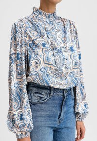 Blusa a maniche lunghe in motivo paisley blu e pesca, con collo alto arricciato e dettagli con bottoni. Abbinata a jeans in denim a vita alta.