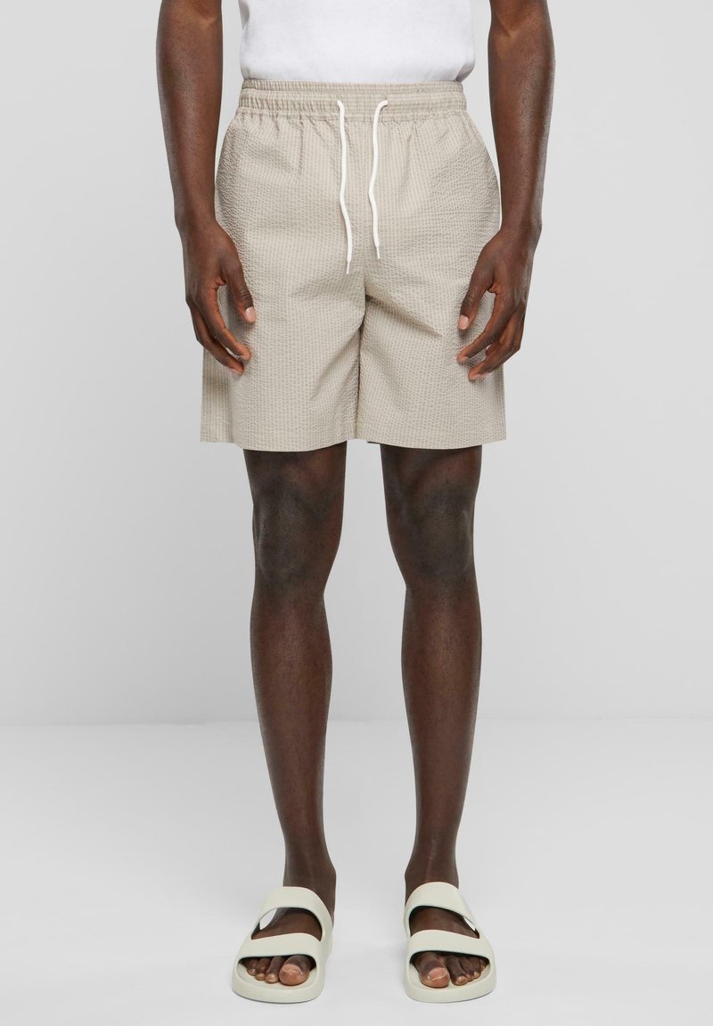 Homme portant un short beige texturé avec des cordons blancs, un t-shirt blanc et des sandales blanches à glissière, debout sur un fond clair uni.