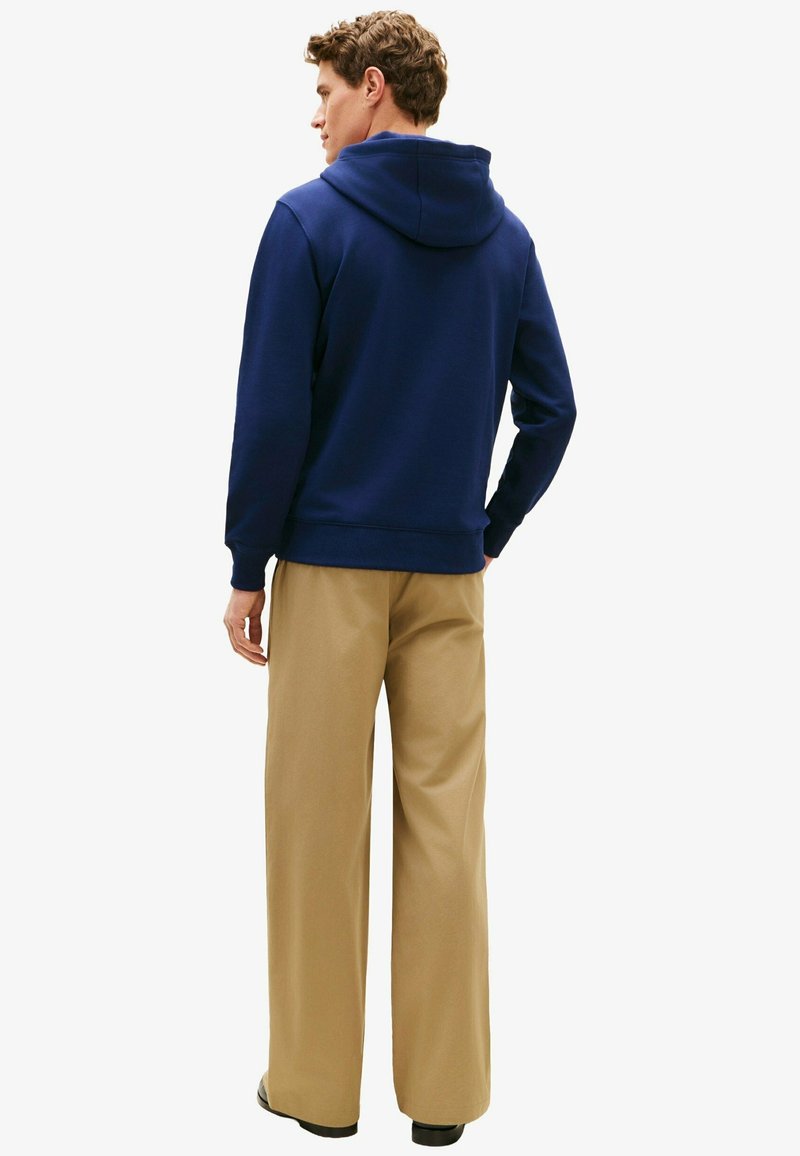 Homme de dos portant un sweat à capuche bleu marine et un pantalon large beige sur un fond blanc uni.