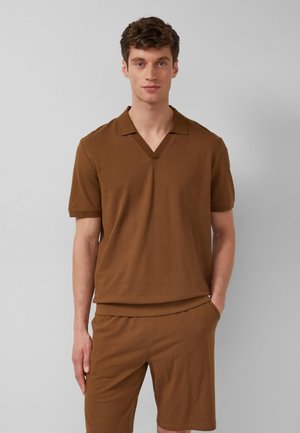 Jeune homme debout, main dans la poche, portant une chemise marron à manches courtes avec col et un short assorti sur un fond gris uni.