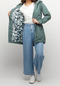 Lekka, zielona wodoodporna kurtka z podszewką w kwiatowy wzór, zestawiona z luźnymi, jasnoniebieskimi spodniami i białymi trampkami. Widać neutralny t-shirt.