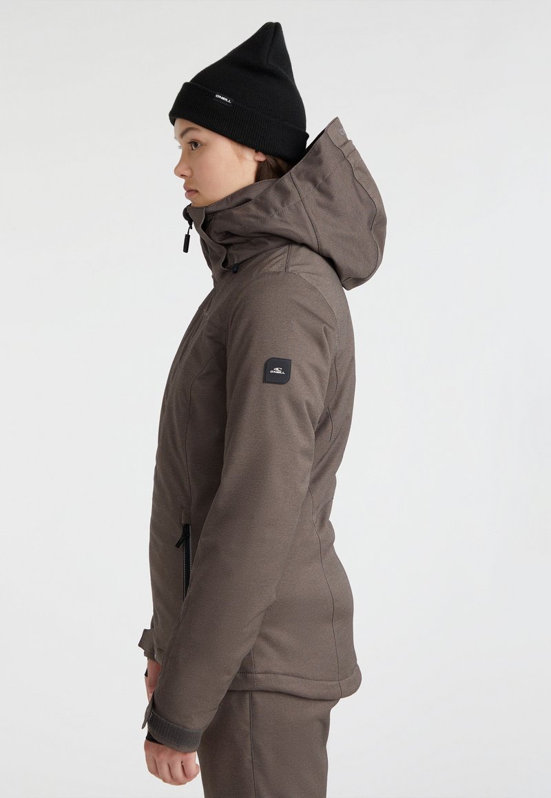 Frau in einer braunen Kapuzenjacke und schwarzer Mütze, die seitlich vor einem schlichten weißen Hintergrund steht.