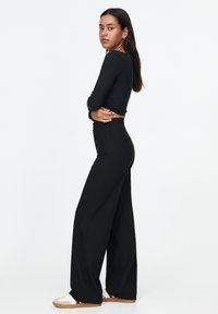 Zwarte lange mouwen top en brede zwarte broek, beide gemaakt van zachte stof. Het model poseert zijwaarts en toont een getailleerde stijl met casual sneakers.
