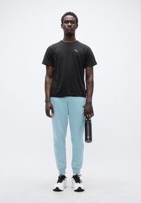 Μαύρο αθλητικό μπλουζάκι, ανοιχτό μπλε φόρμες με ελαστικά τελειώματα και μαύρα αθλητικά παπούτσια. Κρατάει μια μαύρη φιάλη νερού. Απλός σχεδιασμός και χρωματική παλέτα.