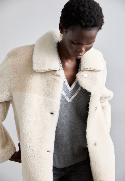 Kermakanvärinen shearling-takki, jossa on suuri kaulus, nappikiinnitys edessä ja harmaa V-kaula-aukkokenkät sisällä. Pehmeä rakenne ja rento istuvuus.