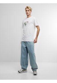 Weißes Baumwoll-T-Shirt mit einem grafischen Design eines Basketballkorbs, umgeben von Blättern. Kombiniert mit hellblauen, locker sitzenden Jeans und silbernen Sneakers.