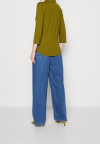 Oliven Grøn, tætsiddende top med trekvartærmer, parret med løse, midtblå, højtaljede jeans. Gule hæle fuldender outfittet.