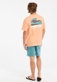 Koraalkleurig T-shirt met korte mouwen en golfafbeelding op de achterkant, gecombineerd met teal shorts. Model staat in slippers op een effen witte achtergrond.