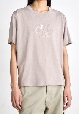 Osoba ubrana w jasnobeżową koszulkę Calvin Klein Jeans z krótkim rękawem i beżowe spodnie na jednolitym białym tle.