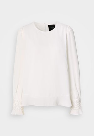 Biała bluzka z długim rękawem, okrągłym dekoltem, plisowanymi ramionami oraz ozdobami z biżuterii przy mankietach, charakteryzująca się gładką, satynową fakturą.