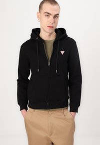 Sweatshirt preto com fecho e capuz ajustável, bolsos frontais e um pequeno logotipo triangular no lado esquerdo do peito. Usado sobre uma camisa verde-oliva.