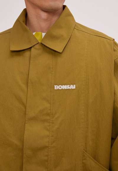 Jachetă verde măslin cu guler mandarin, buzunar lateral și un logo texturat "BONSAI" în alb pe piept. Material ușor cu o finisare netedă.