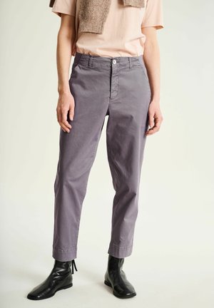 Persona que lleva pantalones grises entallados, botas negras hasta el tobillo, una camisa de manga corta rosa claro y un suéter beige colgado sobre los hombros.