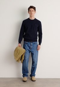 Navy sweater, light blue jeans, beige shoes, holding a light brown jacket. Smooth texture, casual design, standing against a neutral background.