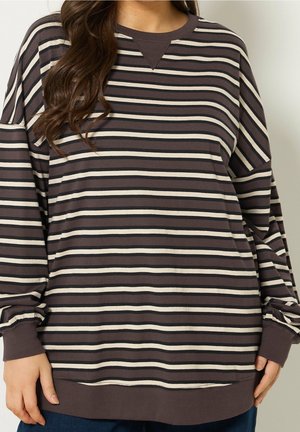 Vrouw draagt een loszittend, lang mouw donkerbruin en beige gestreept shirt met geribbelde manchetten en zoom, staande tegen een effen achtergrond.