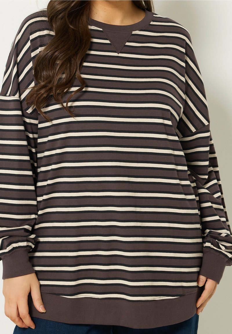 Kobieta ubrana w luźną, długą koszulę w ciemnobrązowe i beżowe paski z prążkowanymi mankietami i dołem, stojąca na jednolitym tle.