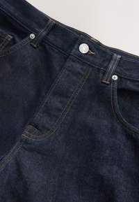 Primo piano di jeans in denim blu scuro che mostrano la tasca anteriore, gli passanti per la cintura e il bottone metallico con dettagli di cucitura visibili.