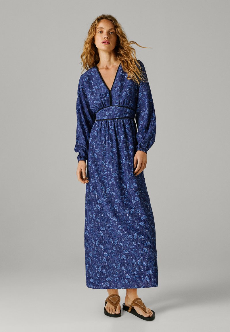 Femme aux longs cheveux bouclés portant une robe maxi bleue à fleurs avec manches longues et des sandales marron sur fond gris.