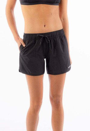 Kobieta ubrana w czarne sportowe szorty z wiązanym pasem i kieszeniami, stojąca z jedną ręką w kieszeni na białym tle.