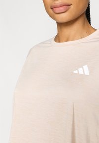 Jasnoróżowa koszulka z okrągłym dekoltem, wykonana z miękkiego materiału. Na lewej stronie klatki piersiowej mały biały trójkątny logo.