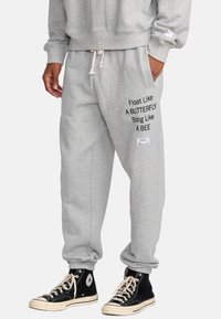 Pantaloni della tuta grigi realizzati in cotone misto, con frasi stampate in nero sulla gamba sinistra e un'etichetta con logo a destra. Polsini elasticizzati alle caviglie.
