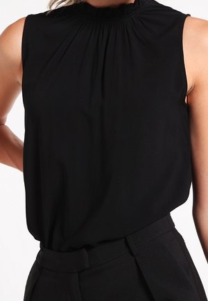 Zwarte mouwloze top met een geplooide hals, gemaakt van zachte stof. Heeft een losse pasvorm en subtiele draping. G gedragen met hoge taille zwarte broek.