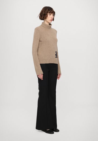 Kaki Rollkragenpullover mit strukturiertem Strick, versehen mit einem kleinen gestickten Design am Saum, kombiniert mit schwarzen weit geschnittenen Hosen.