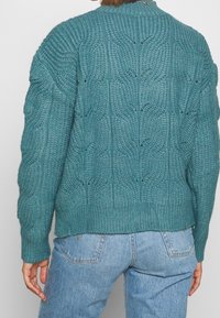 Maglione lavorato a maglia in verde teal con un motivo testurizzato, orlo e polsini a costine, e collo alto. La vista posteriore mostra cuciture strutturate e dettagli ad accesso.