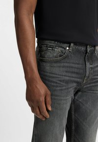 Jeans in denim grigio scuro con una leggera texture sbiadita e cuciture gialle. La vita presenta un'etichetta con il logo 'BOSS' sul lato sinistro.
