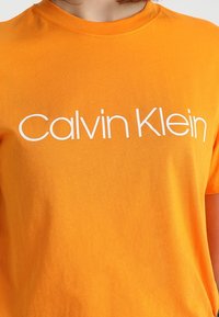 Camiseta de algodón naranja con un logo blanco de "Calvin Klein" en una fuente audaz y moderna en el pecho. Mangas cortas y cuello redondo.