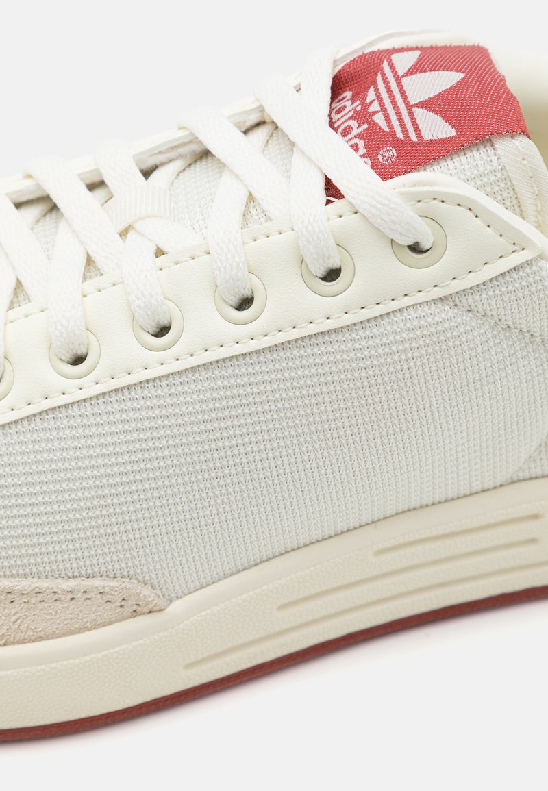 Sneakers blancs avec une tige en tissu texturé, une étiquette de marque rouge bien visible et des accents en suède beige clair. Présente des lacets blancs plats et des œillets.