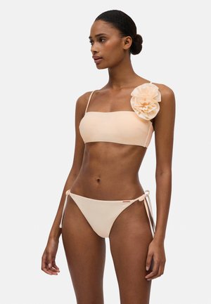 Conjunto de bikini melocotón con top bandeau que destaca por un gran acento floral y braguitas laterales atadas. Textura suave, diseño minimalista.