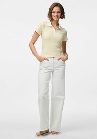 Camisa tipo polo de manga corta con rayas amarillas y blancas, combinada con jeans blancos de pierna recta. El modelo está de pie con las manos en los bolsillos.