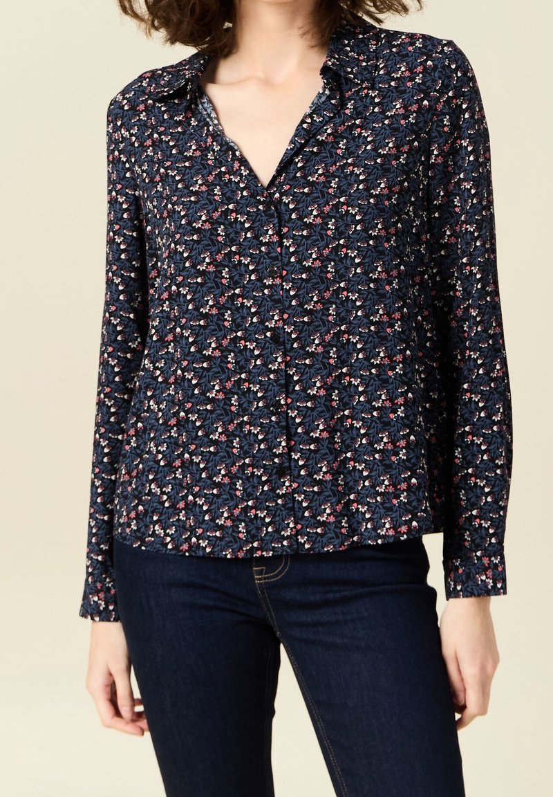 Femme portant un chemisier bleu marine avec de petits motifs floraux roses et blancs, associé à un jean bleu foncé sur un fond uni.