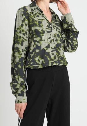 Femme portant une chemise boutonnée à manches longues, à motifs verts et noirs, associée à un pantalon noir avec des bandes blanches sur les côtés.