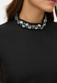 Černý top s výrazným límcem zdobeným průhlednými oválnými krystaly a kovovými akcenty, který nabízí hladkou texturu.