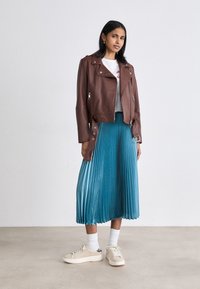 Woman wearing a brown leather jacket, white graphic tee, blue pleated midi skirt, white socks, and beige sneakers standing on a plain floor.